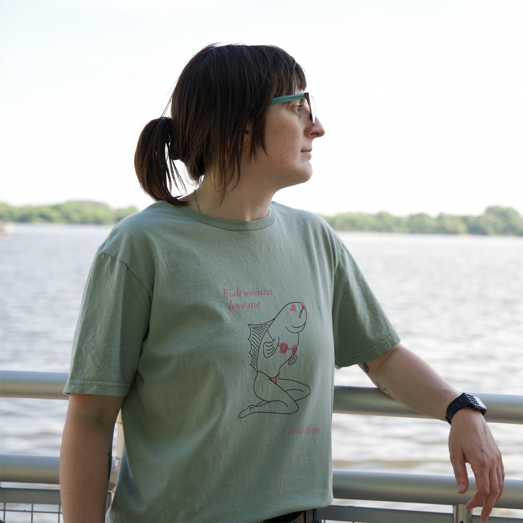 Model wearing Sirena (Fish Woman) T-Shirt from Cryptid Records in sage green, standing at a riverside railing